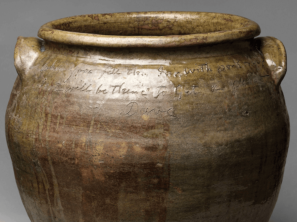 In this photo, an alkaline glazed storage jar created by a potter named Dave sits against a gray background in a museum. The overhead light hits the jar in a way that illuminates his handwriting and signature.