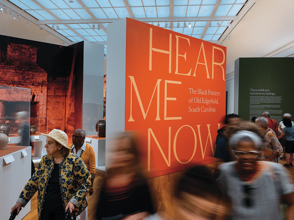A crowd moves through the University of Michigan Museum of Art’s exhibition space in this photo. In large yellow lettering behind the crowd, Hear Me Now: The Black Potters of Old Edgefield, South Carolina is in bold type on an orange wall.  